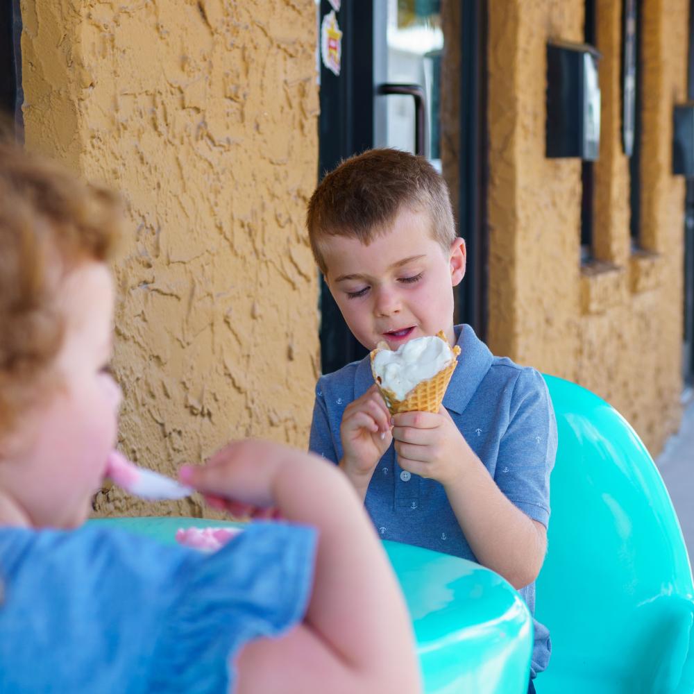 Two young children sit outside on bright turquoise chairs, enjoying ice cream at a table in front of a shop in Downtown St. Cloud, Florida. One child licks a waffle cone while the other eats with a spoon.