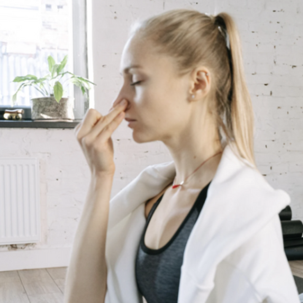 Two people sit cross-legged in a bright, minimalist studio practicing alternate nostril breathing as part of a mindfulness or yoga session.