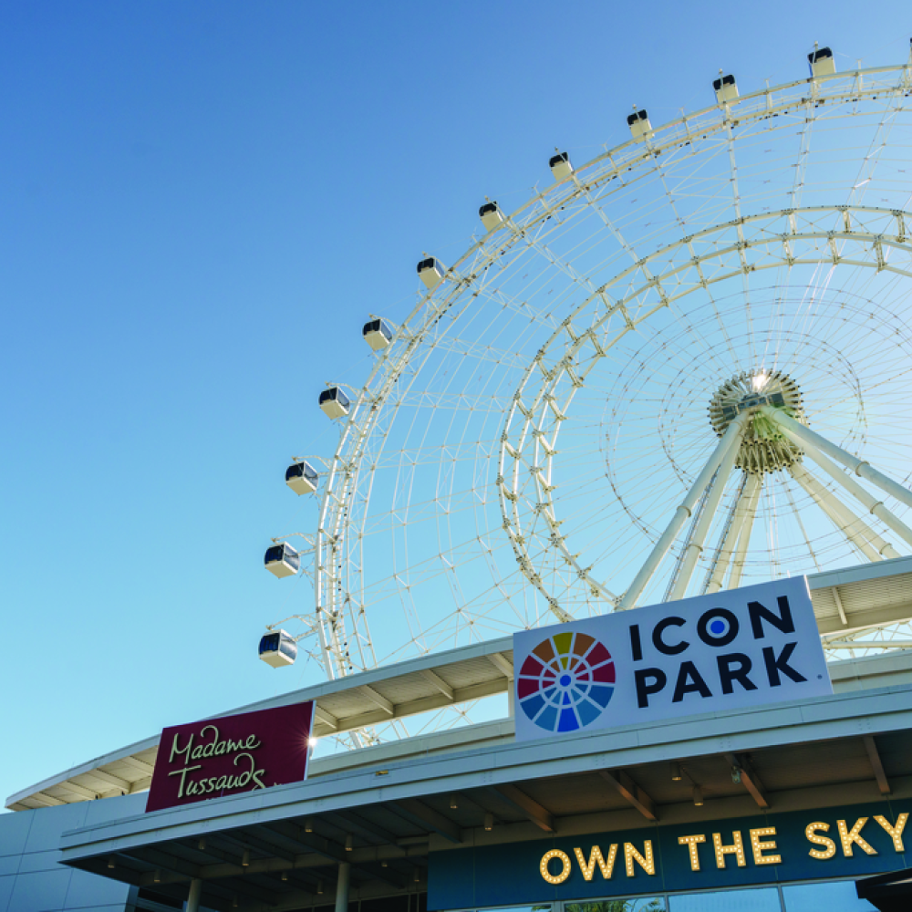 A low-angle view of The Wheel at ICON Park in Orlando, with clear blue sky overhead and signs for ICON Park and Madame Tussauds visible at the entrance.