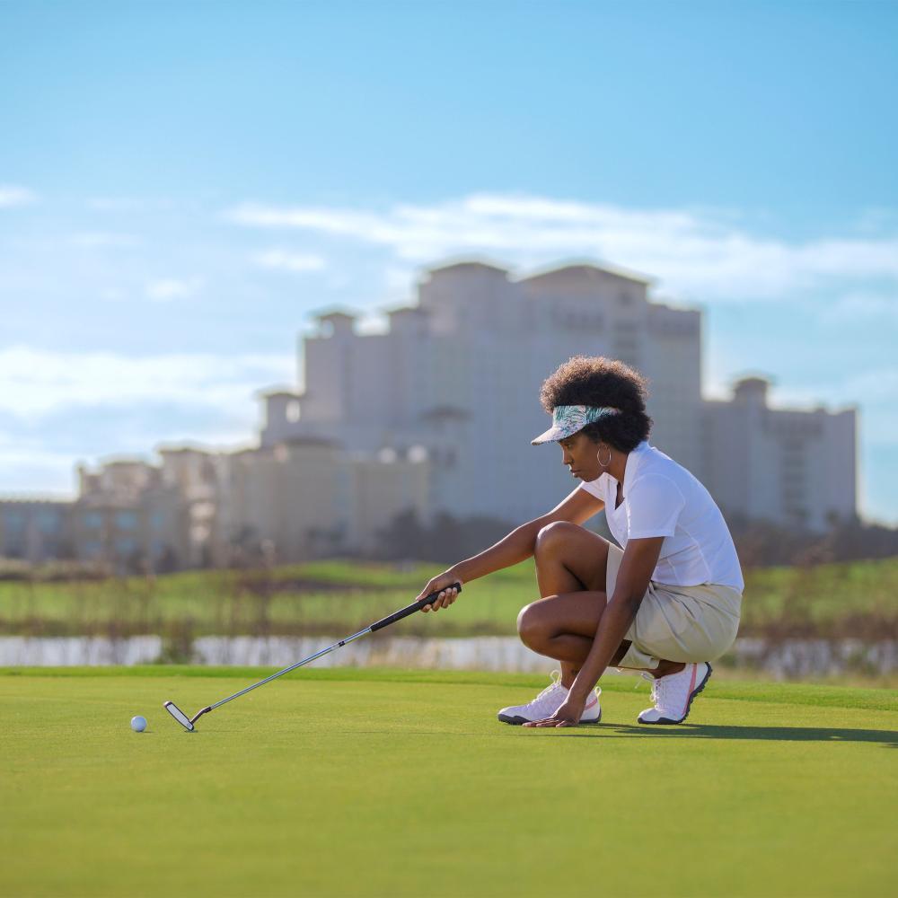 Woman golfer lines up a putt on a sunny day at the Omni Orlando Resort at ChampionsGate in Kissimmee, Florida, with the resort’s luxury hotel visible in the background.
