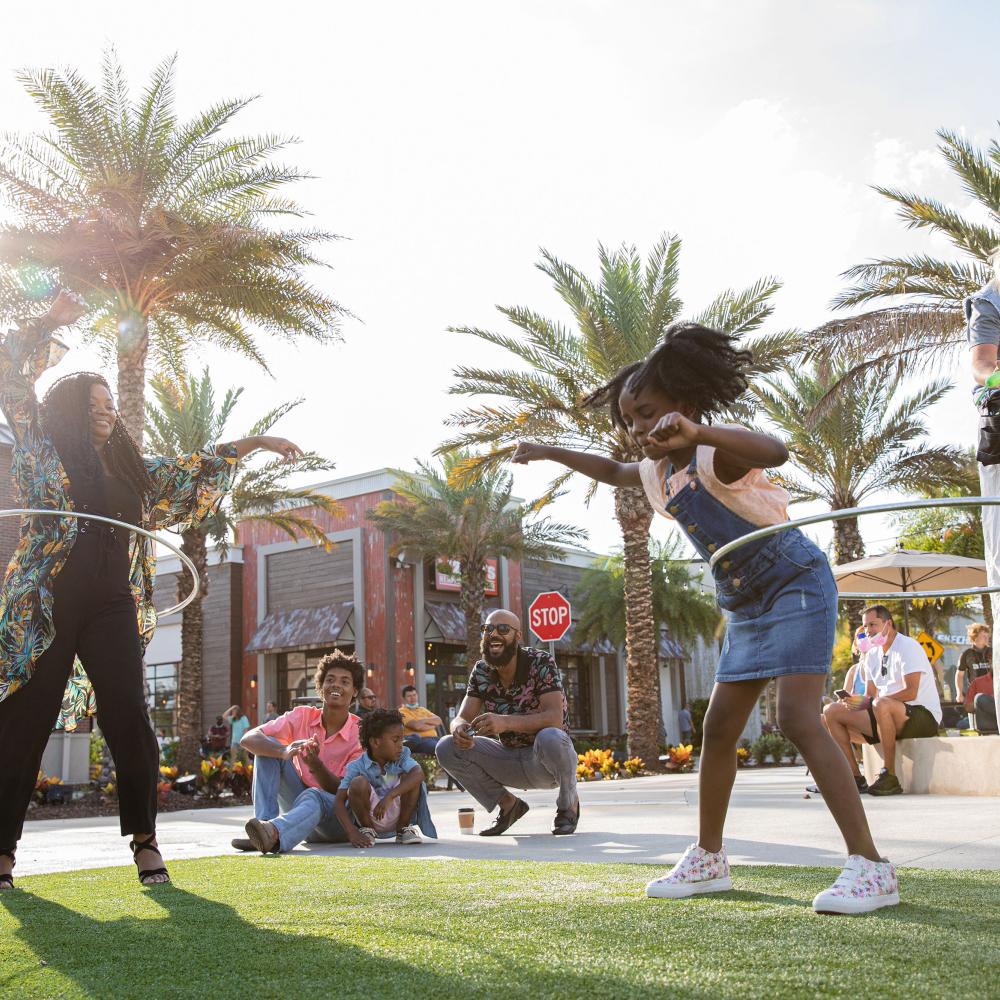 Families enjoying outdoor entertainment at Promenade at Sunset Walk in Kissimmee, Florida, with a woman and young girl hula-hooping under palm trees on a sunny afternoon.