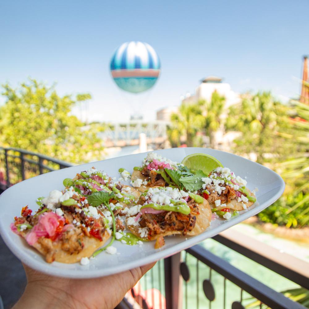 A plate of carnitas tostadas garnished with lime, cilantro, and queso fresco at Planet Hollywood in Disney Springs, with the Aerophile balloon visible in the background.