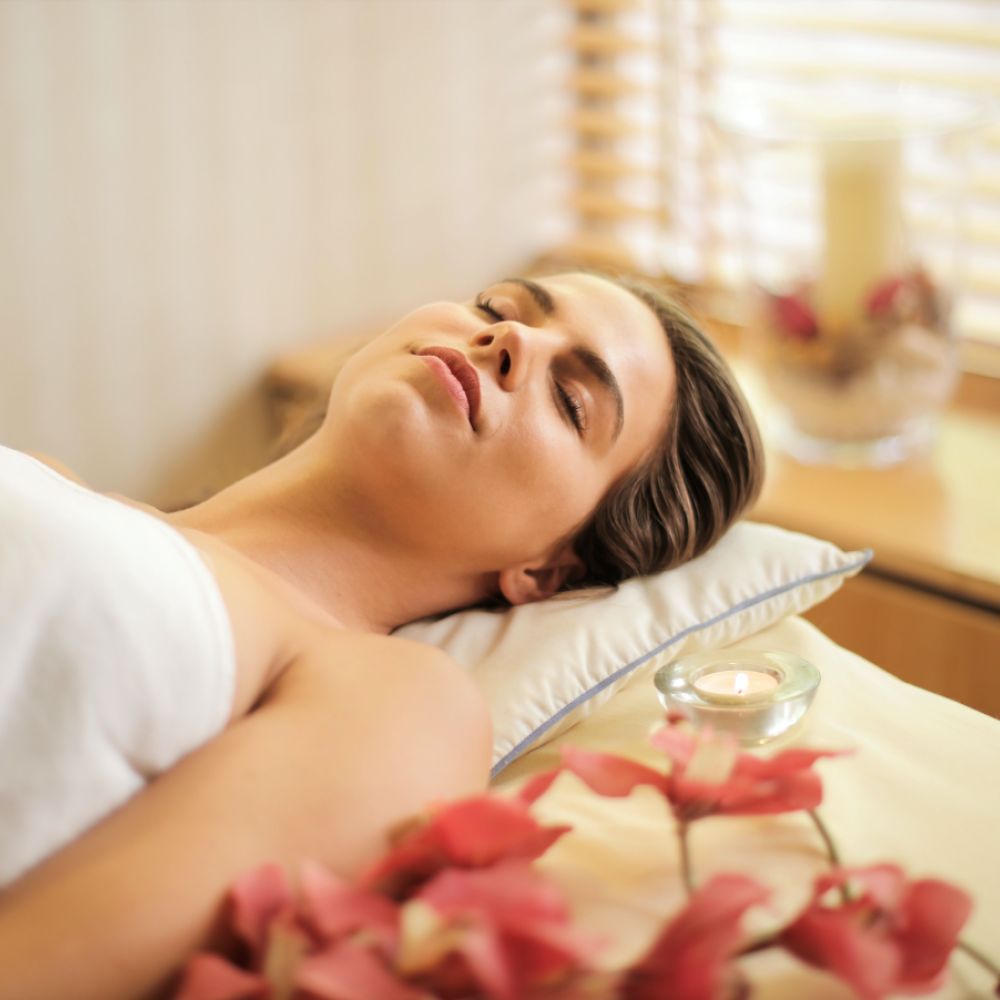 Woman relaxing on a massage table wrapped in a white towel, surrounded by soft lighting, flowers, and candles in a tranquil spa setting.