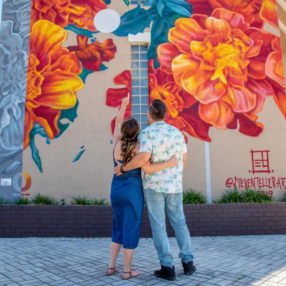 A couple stands arm in arm, looking up and pointing at a large, colorful floral mural painted on the side of a building in a sunny downtown setting.