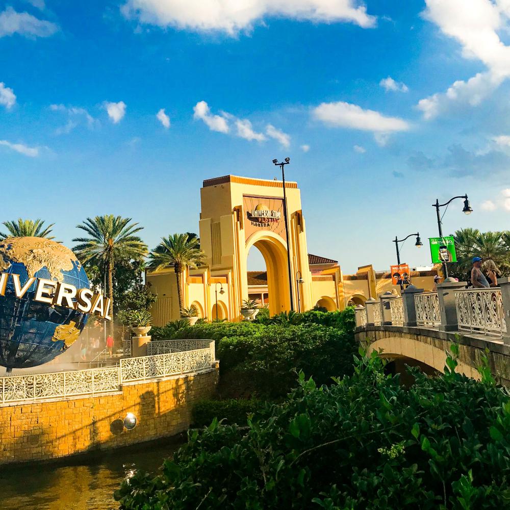 The iconic Universal Studios globe and entrance archway sit under a bright blue sky, with visitors walking across a nearby bridge lined with banners and palm trees.