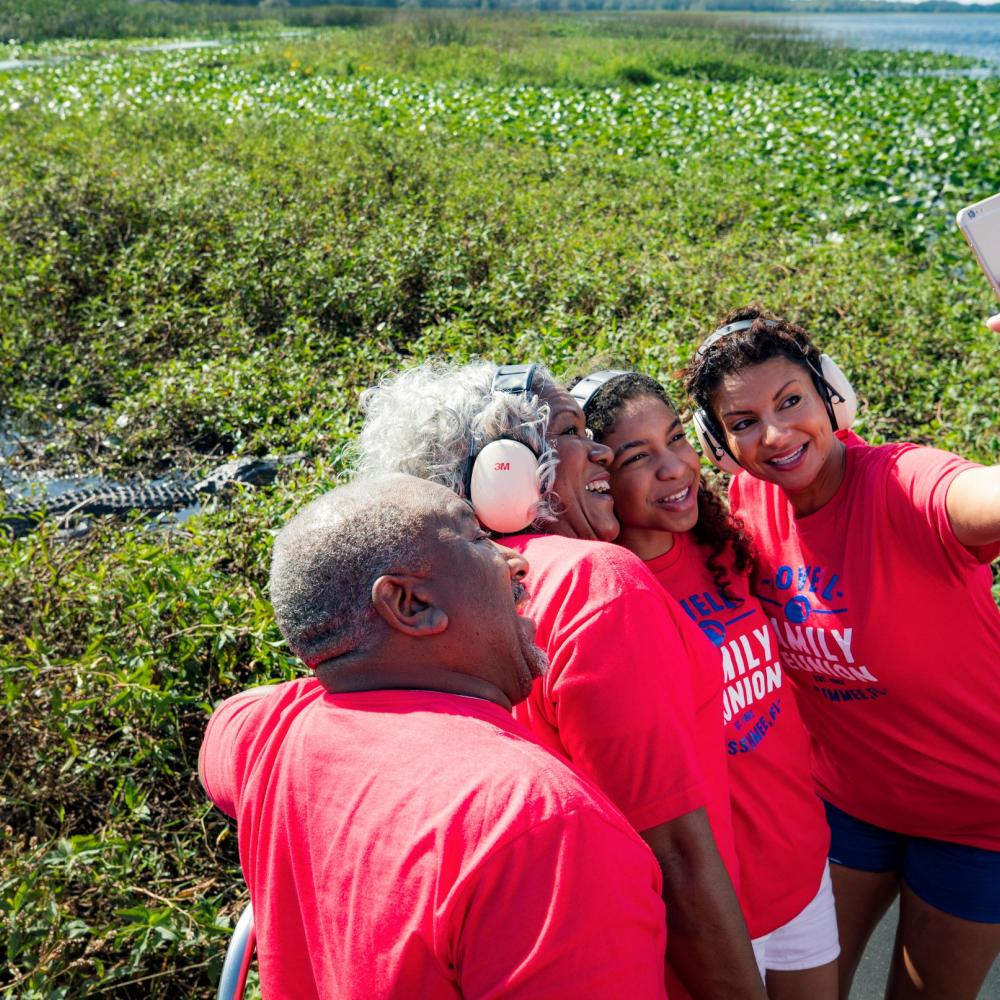 A multigenerational family wearing protective headsets takes a smiling selfie on an airboat ride through a marsh, with lush wetlands and an alligator visible in the background.