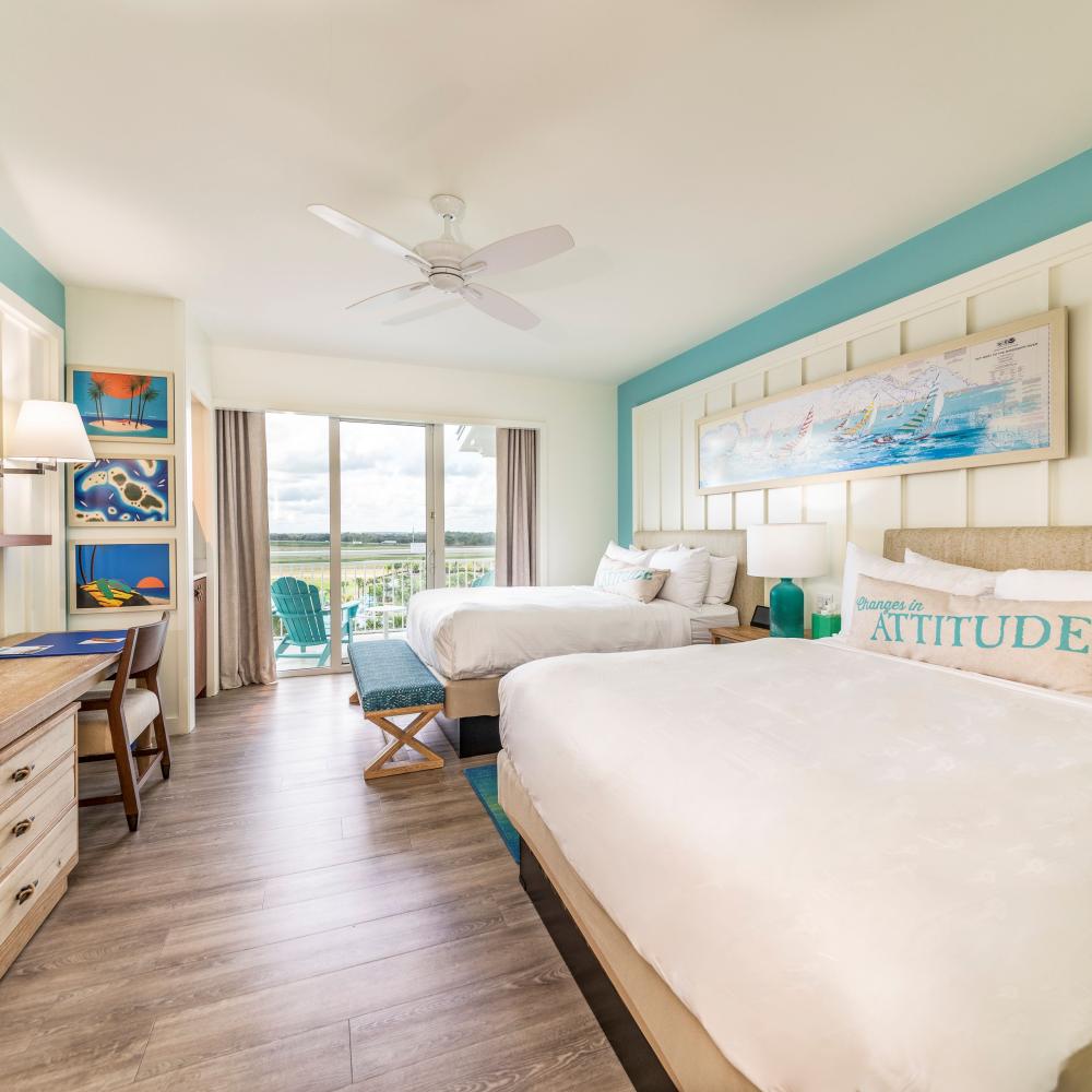 A bright, coastal-themed hotel room with two queen beds, colorful artwork, a desk, a mounted TV, and sliding glass doors leading to a balcony with outdoor seating and scenic views.