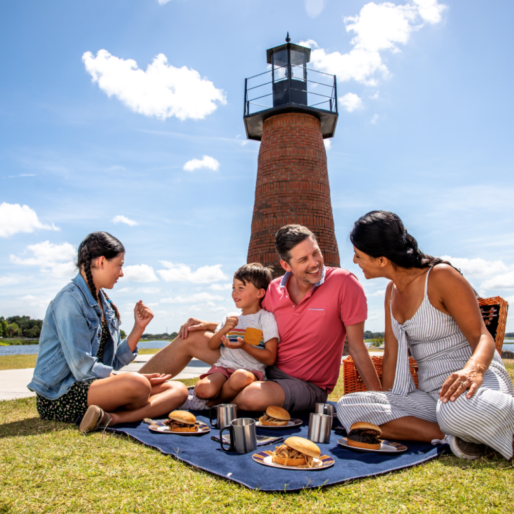 A family enjoying a picnic on a blanket near a brick lighthouse at a waterfront park, sharing food and conversation under a bright blue sky.