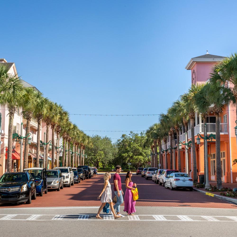 Friends crossing a palm-lined street in Celebration, Florida, surrounded by colorful pastel buildings, parked cars, and clear blue skies.