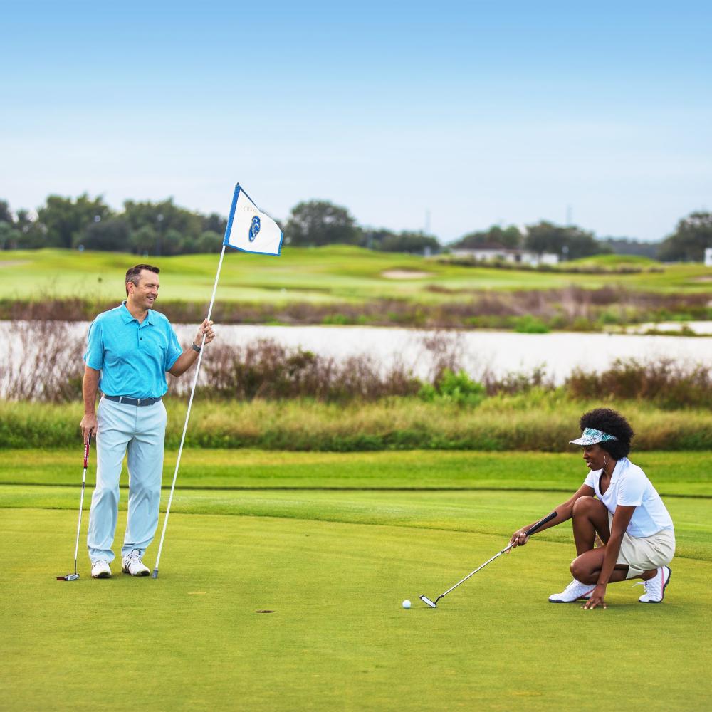 Two golfers on a putting green at a scenic Kissimmee golf course, with one person preparing a putt and another holding the flag.