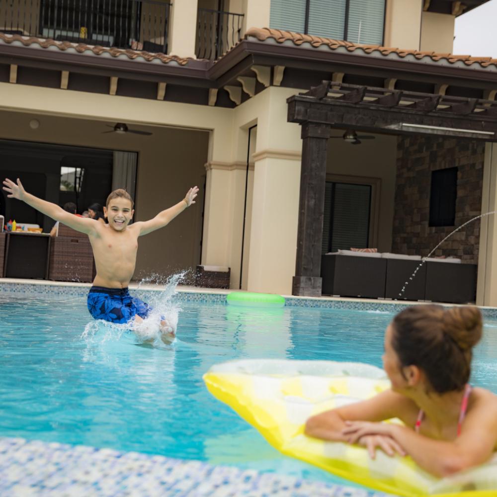 A young boy joyfully jumps into a private vacation home pool while a woman relaxes on a yellow float, smiling toward him. The luxury home’s patio and outdoor seating area sit in the background.