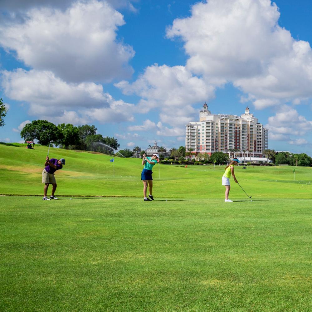 Group of people practicing their golf swings on a sunny day at Reunion Resort’s golf course in Kissimmee, Florida, with the resort’s main tower visible in the background.