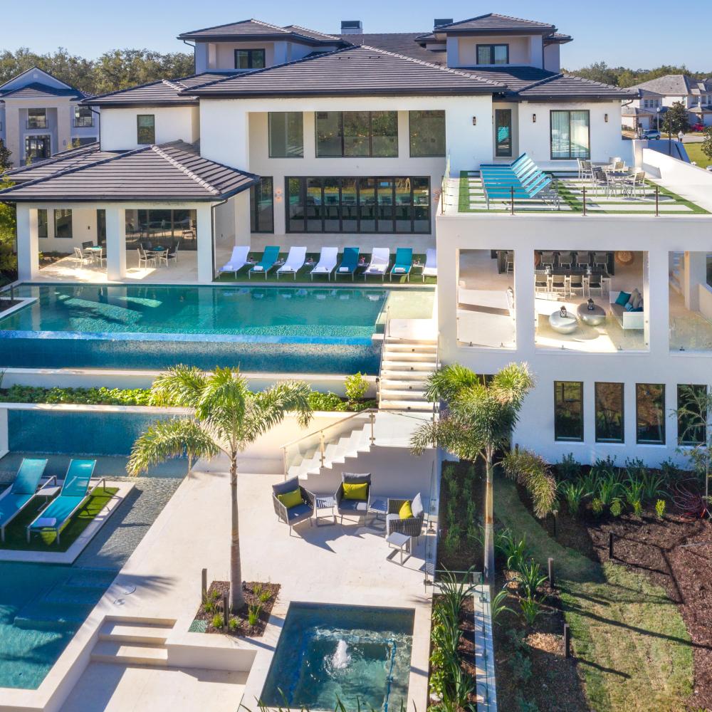 A large modern vacation home with multiple outdoor pools, sun loungers, terraces, and tropical landscaping, viewed from above in a residential resort community.