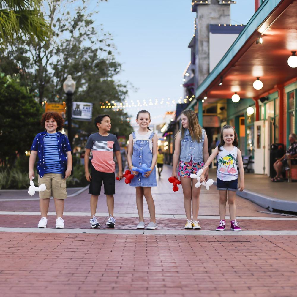 A group of six children stand side by side on a brick street at Old Town Kissimmee in the evening, holding balloon animals with string lights and colorful storefronts behind them.