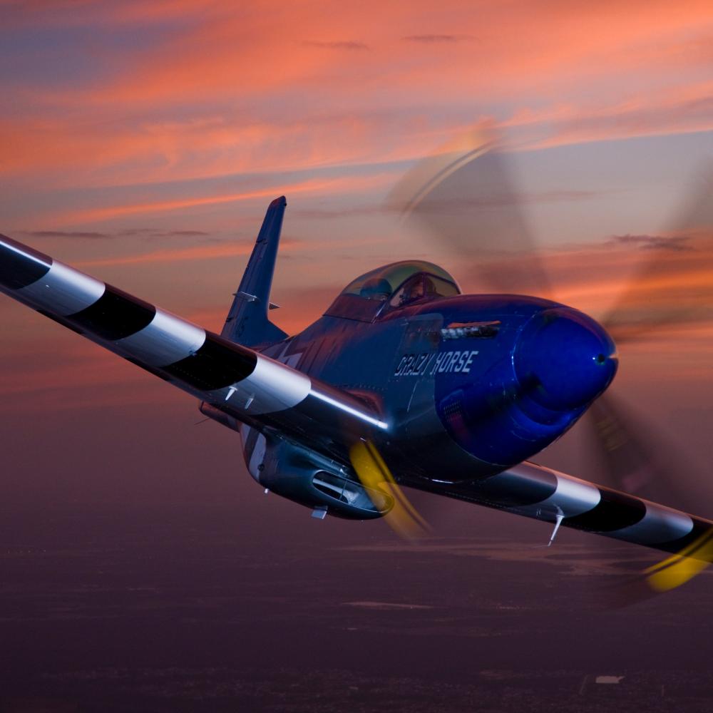 Blue P-51 Mustang aircraft named “Crazy Horse” soars through the sky at sunset, its propeller in motion against a vivid orange and purple horizon.