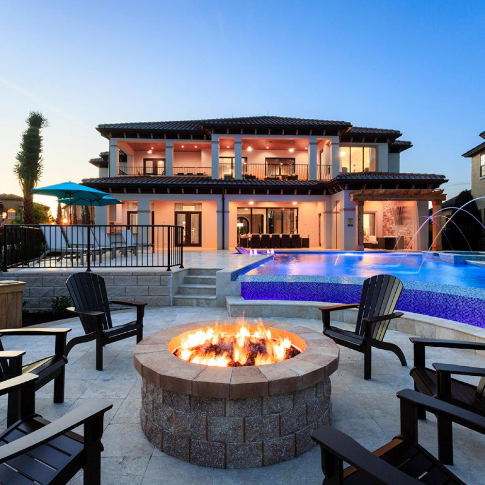 A two story Mediterranean style vacation home sits in the back ground, with a pool, chairs and fire pit in the foreground at dusk.