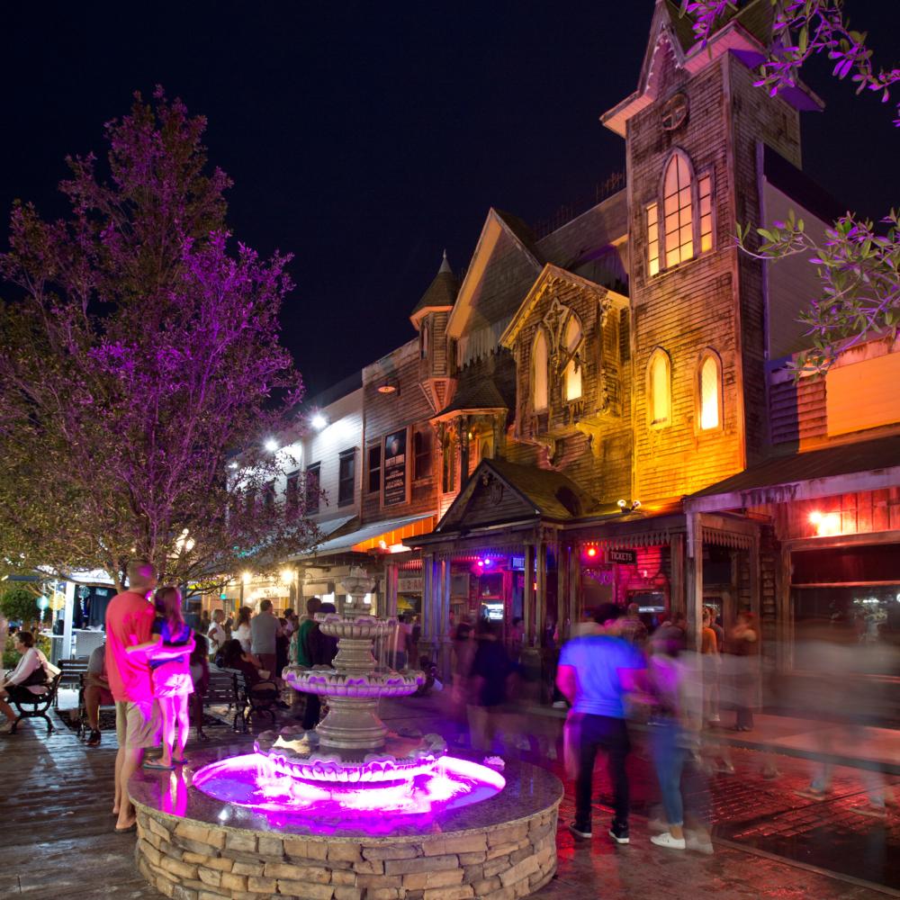 Nighttime scene at Old Town Kissimmee with visitors walking past the illuminated Mortem Manor haunted attraction and a glowing purple fountain in the foreground.