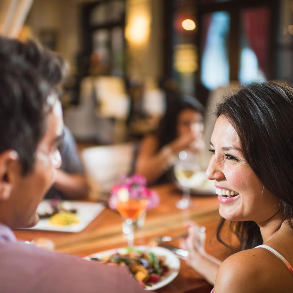 A woman smiles at her companion as they sit around a communal table to enjoy a delicious meal.