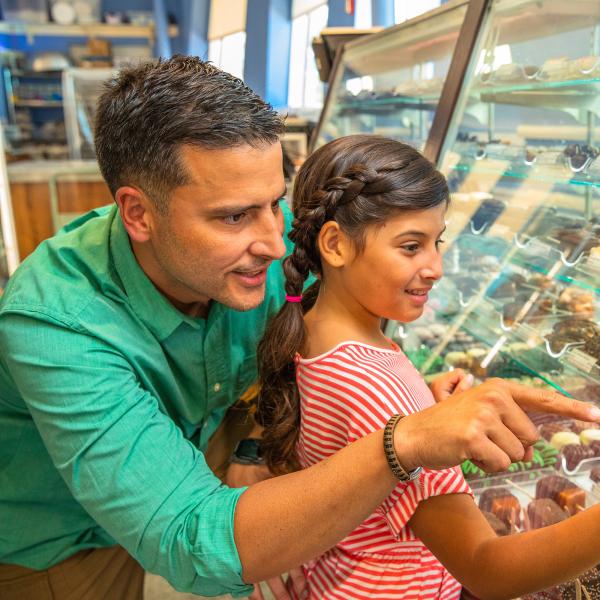 A father and daughter smile and point at assorted chocolates and sweets inside a glass display case at a candy shop.