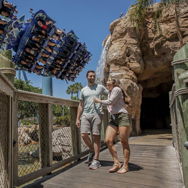 A couple walks across a wooden bridge at SeaWorld as a roller coaster full of riders swoops overhead.