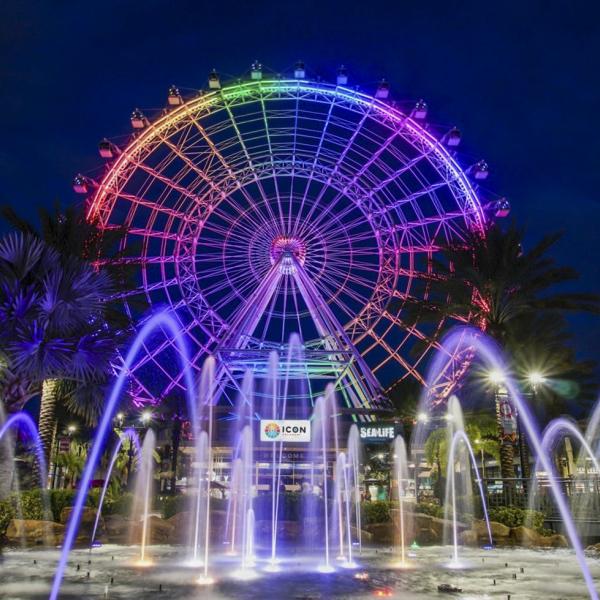 Nighttime view of The Wheel at ICON Park in Orlando, glowing with rainbow lights above a fountain display.