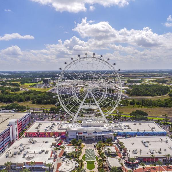 Aerial view of the Orlando Observation Wheel at ICON Park, surrounded by shops, restaurants, parking garages, and wide stretches of green landscape under a bright, partly cloudy sky.