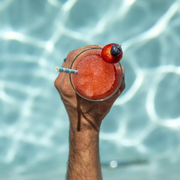 A hand holds a bright red frozen drink garnished with a strawberry and blueberry by the edge of a shimmering blue pool.