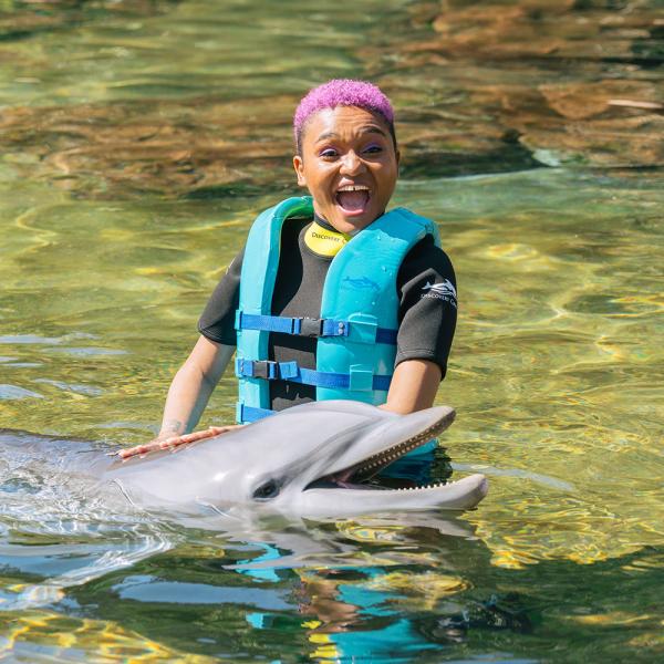 A smiling woman wearing a wetsuit and turquoise life vest interacts joyfully with a dolphin in the clear waters of Discovery Cove.