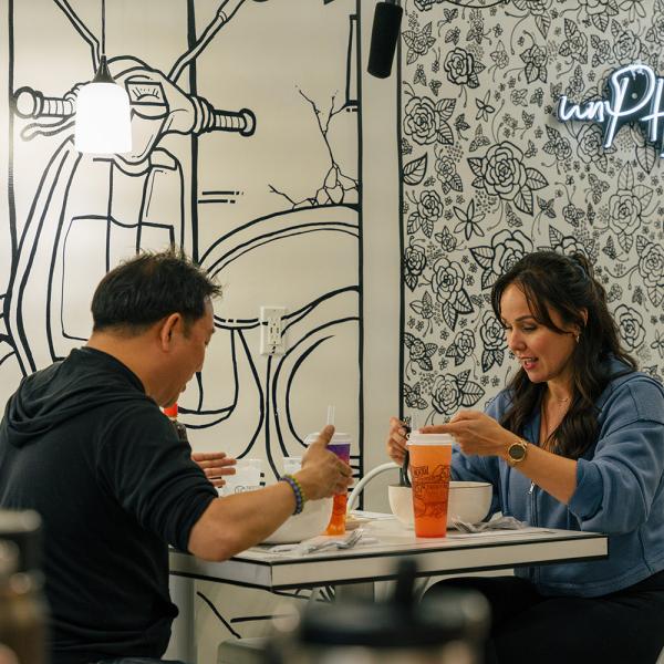 Two people sit at a small table inside a pho restaurant, eating from large bowls of soup with iced drinks beside them. The walls feature black-and-white floral and scooter illustrations, along with a neon sign that reads “be unPHOgettable.”