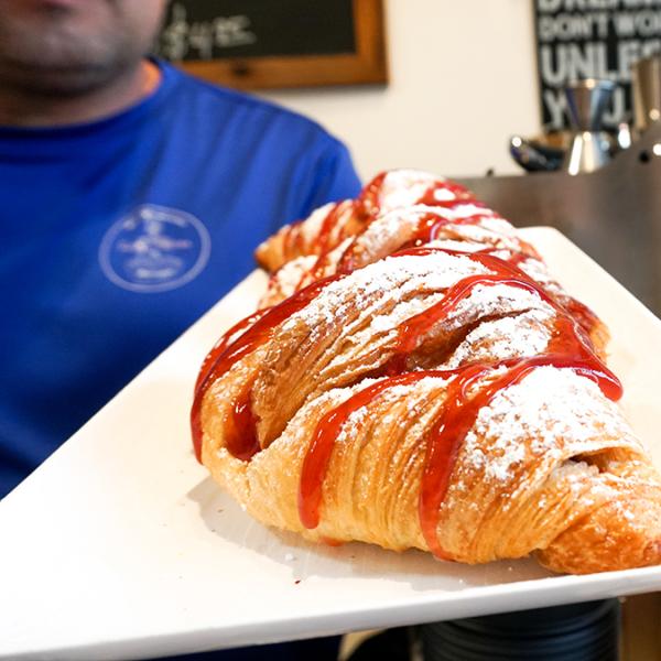 A powdered sugar–dusted croissant drizzled with strawberry sauce is served on a white plate inside a cozy café, with espresso equipment in the background.