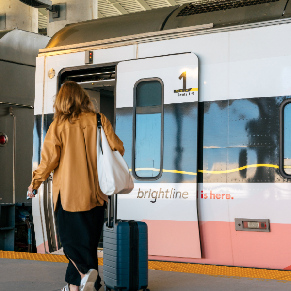 A traveler with a rolling suitcase boards a Brightline train at the station, walking toward the open door.