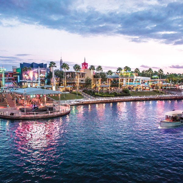 Evening view of Universal CityWalk in Orlando, Florida, with colorful lights reflecting on the water and a boat cruising along the canal near restaurants and entertainment venues.