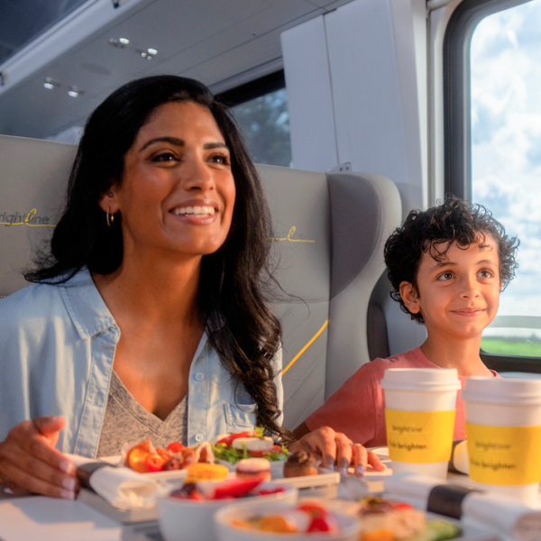 A mother and her young son enjoy a meal together while riding the Brightline train, with sunlight streaming through the window and a scenic view outside.
