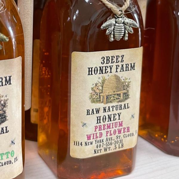 Bottles of raw natural honey from 3Beez Honey Farm in St. Cloud, Florida, displayed on a shelf with decorative bee charms tied around the tops.