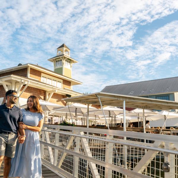 A couple walks hand in hand along the dock outside The BOATHOUSE at Disney Springs, smiling under a bright, partly cloudy sky with the restaurant’s tower and outdoor umbrellas in the background.