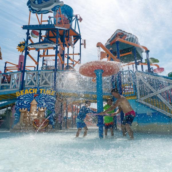 Father with two children getting splashed under a mushroom-shaped water feature at Island H2O Water Park in Kissimmee, Florida, with water slides and a “beach time” sign visible in the background.