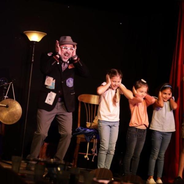 A performer onstage at a family-friendly comedy and magic show stands alongside a group of children, all playfully covering their ears as part of the act.