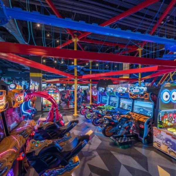 Colorful arcade filled with racing games, neon lights, and an overhead ropes course structure at an indoor entertainment center.