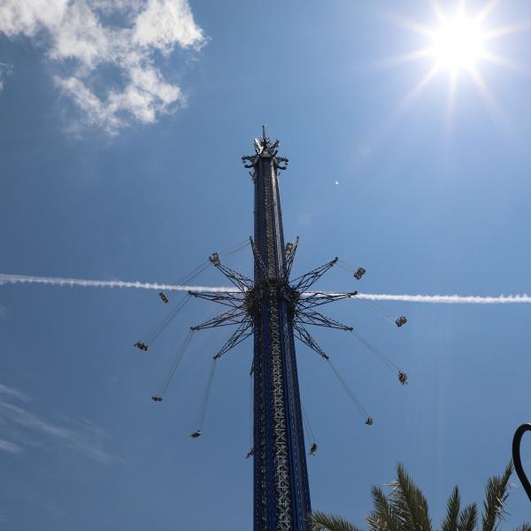 A tall swing ride spins riders high in the air against a bright blue sky, with the sun shining overhead and a jet trail crossing behind the ride.