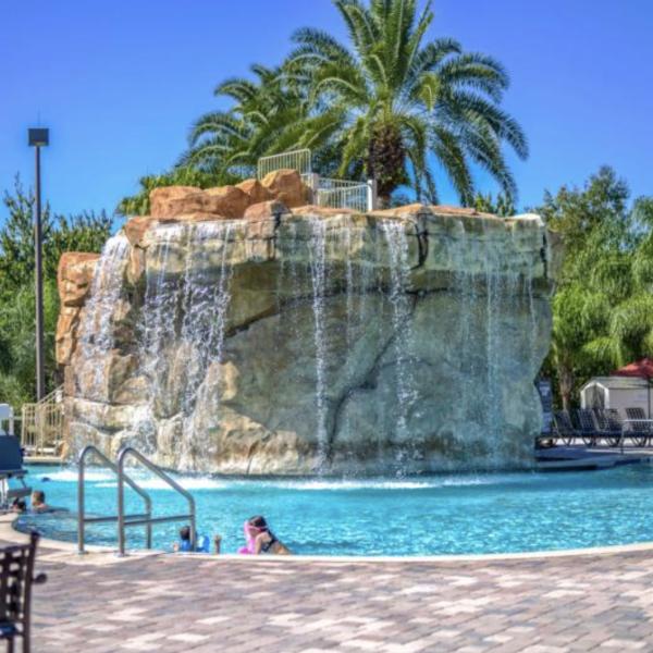 Resort pool featuring a large rock waterfall cascading into a clear blue swimming area, surrounded by palm trees, lounge chairs, and umbrellas.