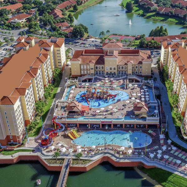 Aerial view of a large resort complex featuring twin hotel towers, a central pool and water play area, and surrounding lakes and palm-lined grounds.