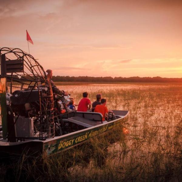A group of people ride a Boggy Creek airboat through tall marsh grasses at sunset, watching the sun dip below the horizon.
