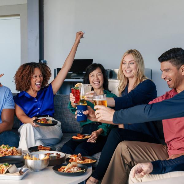 A group of adults sits together sharing food and drinks, laughing and raising their glasses in a celebratory toast.