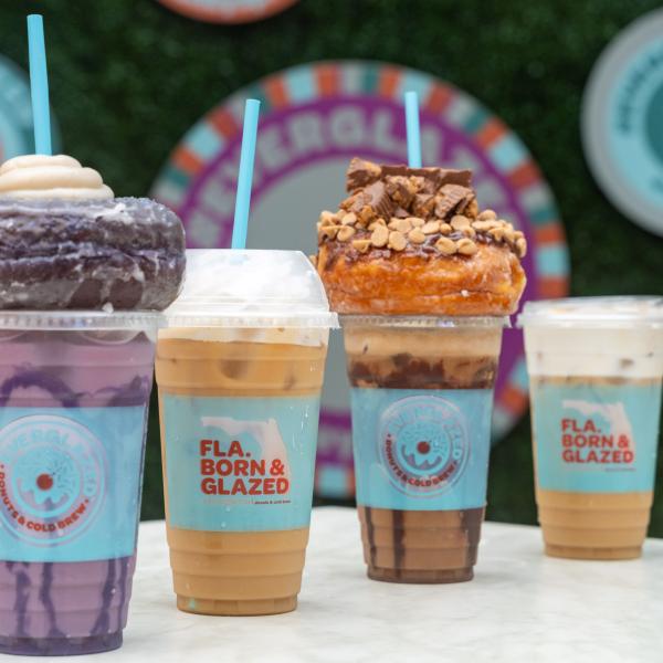 Four drinks are on a white tabletop with greenery in the background. The drink in the front is ube flavored, and it is light purple with dark purple swirls. It is topped with a glazed purple ube doughnut and white frosting. The drink next to it is a conventional cold brew with milk and topped with whipped cream. The next drink is peanut butter flavored and is light brown with chocolate swirls. It is topped with a glazed doughtnut with peanut butter cup crumbles. The last drink is another conventional brew.