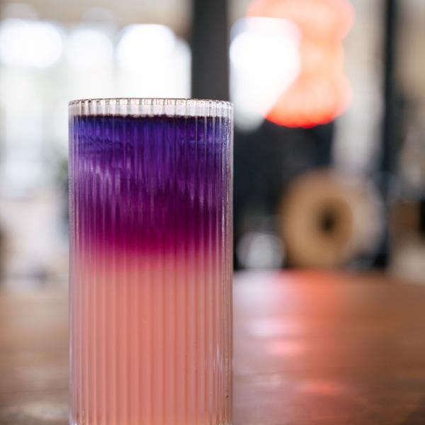 The galaxy lemonade sits in a tall fluted glass on a wooden table. It is light pink on the bottom and it blends into magenta and purple before reaching the blue pea flower tea at the top. It shimmers with edible glitter.
