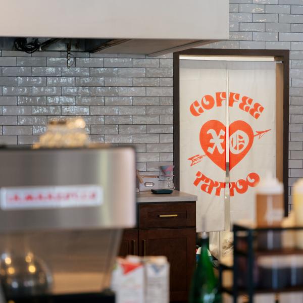 A shot of the interior of XO Coffee shows various coffee making tools and ingredients. In the background, there is a two paneled beige canvas curtain to the kitchen with XO Coffee and Tattoo in bright red letters.