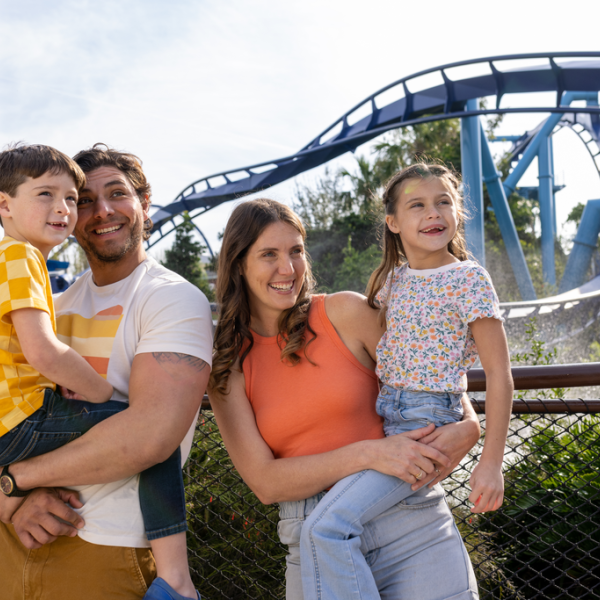 A family of four smiles and poses together in front of a looping roller coaster at a theme park, with parents holding their two young children.