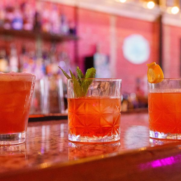 Three vibrant cocktails on the bar at 1881 Restaurant & Bar in Downtown Kissimmee, each amber in hue, served in short glasses, and with garnishes ranging from orange peels, mint, and more.