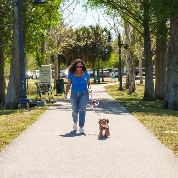 A woman walking her small brown dog on a sunny day at Dan Tarrell Memorial Point in St. Cloud, Florida, with trees lining the paved path.