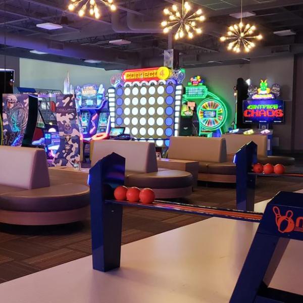 An indoor arcade featuring neon-lit games and a mini bowling lane with orange bowling balls in the foreground.